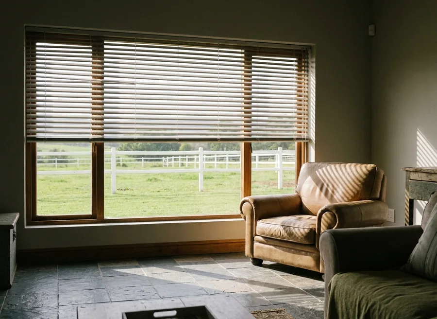 Champagne-toned venetian blind mounted across a wide timber-framed lounge window on a Chartwell-area smallholding, slats tilted so morning light stripes a leather armchair, white paddock railing beyond the glass