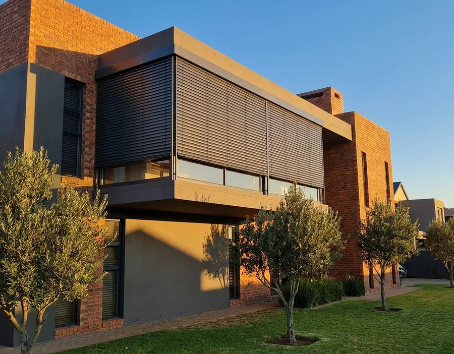 Graphite external venetian blinds in guide rails outside the wide first-floor window of a face-brick Chartwell-area home, slats angled against the low golden-hour sun above a row of olive trees
