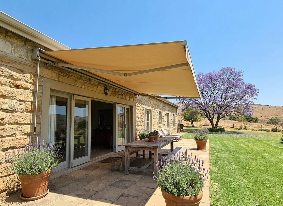 Sand-coloured folding-arm awning mounted above wide patio doors on a Chartwell-area stone house, arms fully extended to shade a terrace table ringed by pot-grown lavender, jacaranda in bloom beyond