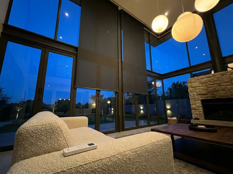 Two motorised charcoal roller blinds halfway down a double-volume window wall in a Chartwell-area home at twilight, the remote lying on the arm of the sofa