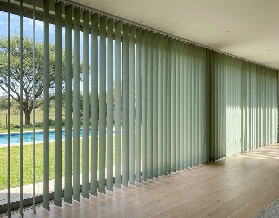 Sage-green vertical blind vanes on a slim ceiling track spanning a wide glass wall in a Chartwell-area home, angled half-open toward the lawn, pool and thorn tree outside