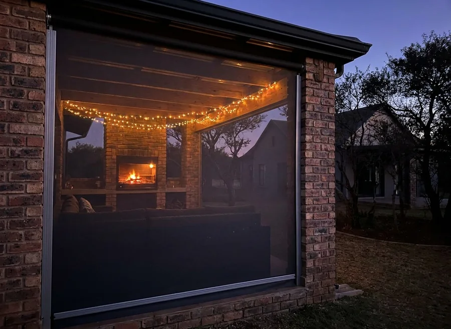 Charcoal mesh zip screen lowered in its side channels between the brick columns of a Chartwell-area braai patio at dusk, string lights and the fire glowing warmly behind the taut fabric